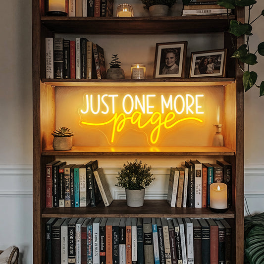 Just One More Page neon sign glowing warm white and yellow on a wooden bookshelf in a cozy home library